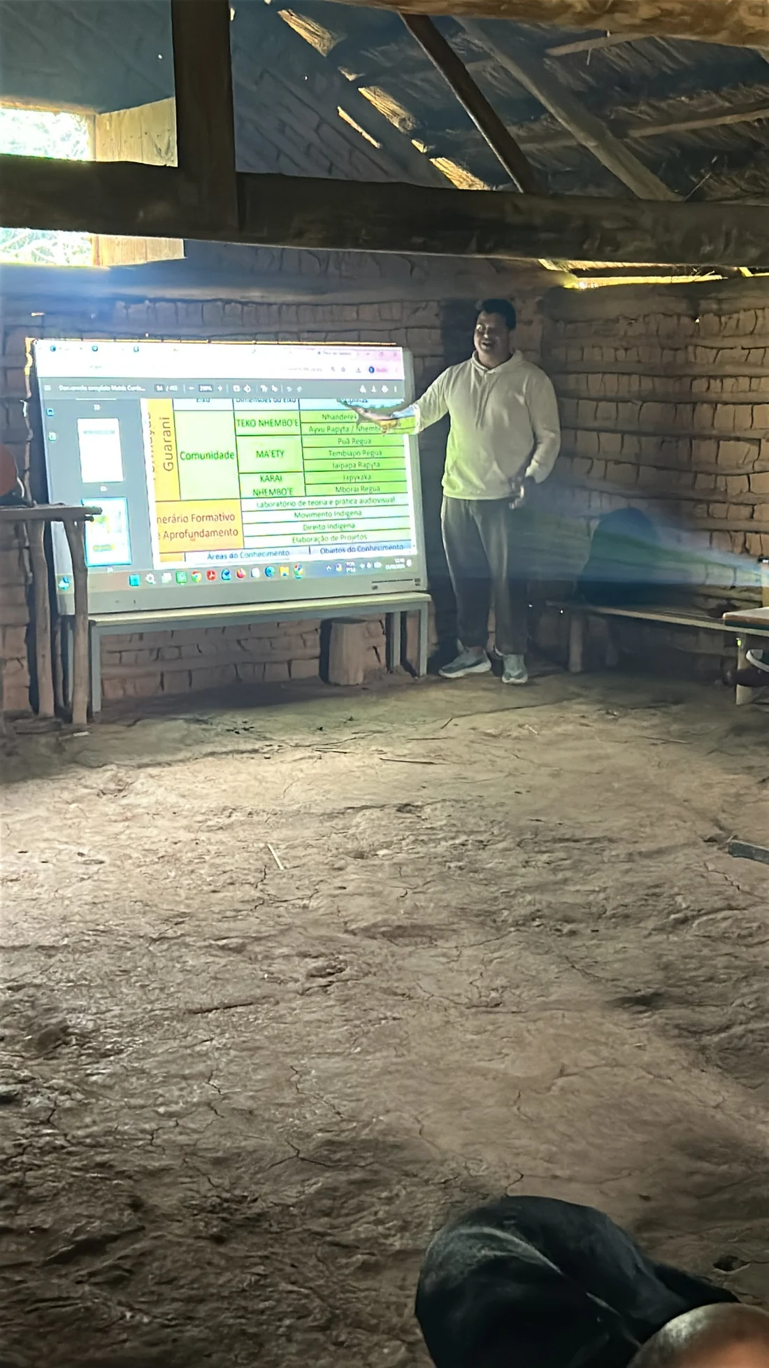 Homem apresenta um quadro projetado numa tela, dentro de uma construção de barro