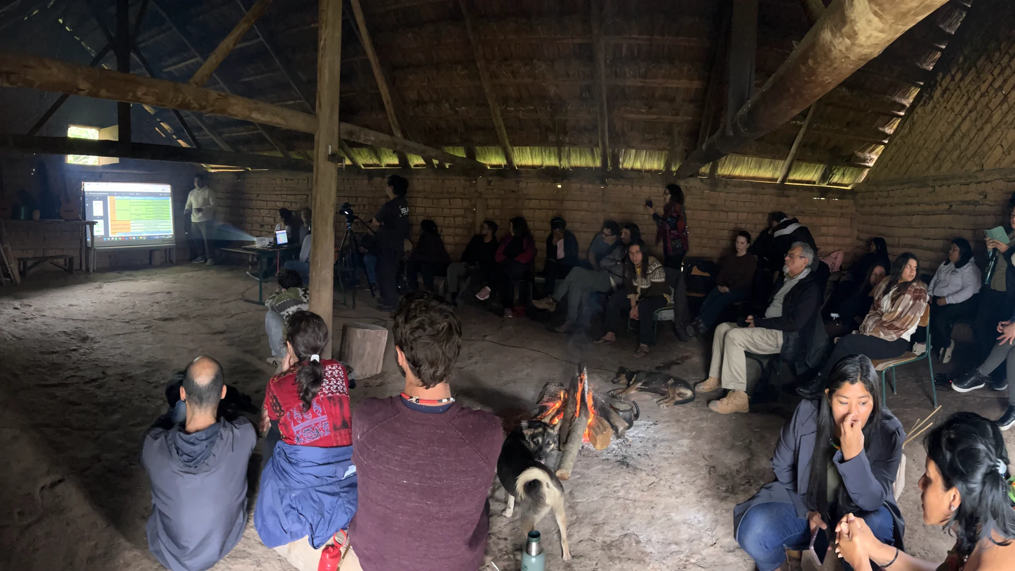 Pessoas sentadas em roda numa construção de barro e madeira, com uma fogueira no chão e uma apresentação projetada na parede